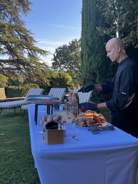 Homme préparant un banquet en plein air sur une table, avec des arbres en arrière-plan.
